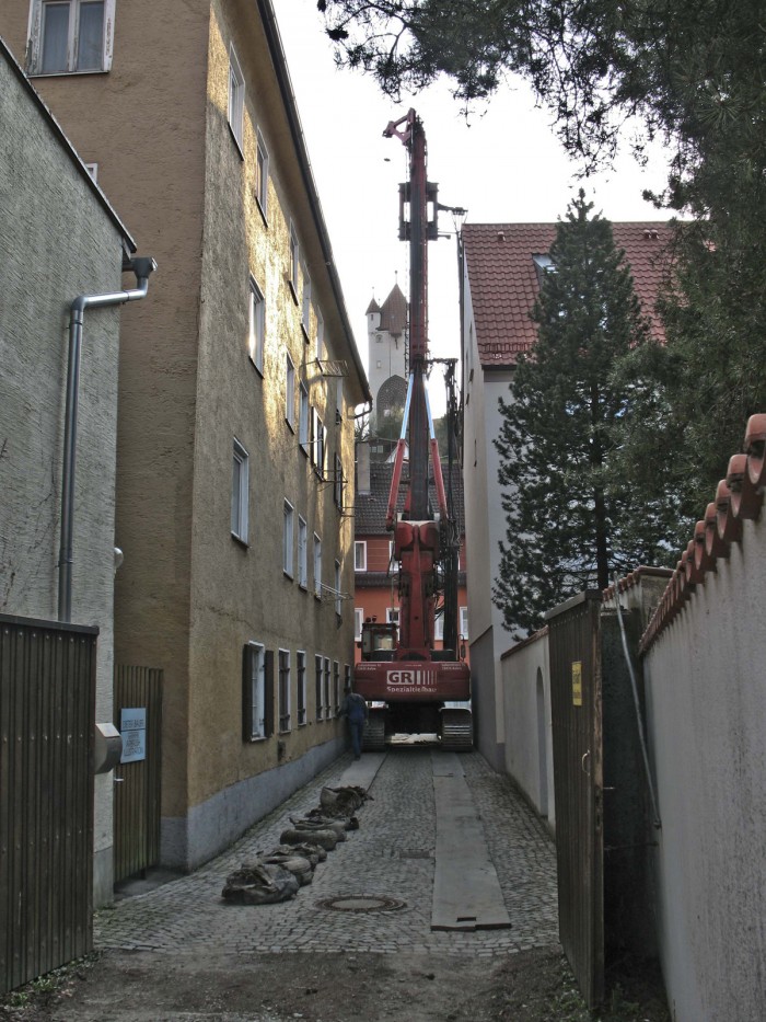 Ein Baustellenfahrzeug mit einem riesigen Bohrer fährt durch eine sehr schmale Gasse zwischen zwei Häuser Richtung Museumshof. Im Hintergrund klein der Fünfknopfturm