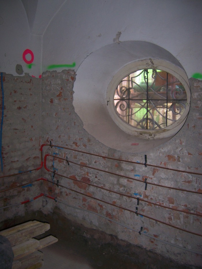 Blick auf eine Wand mit rundem, vergittertem Fenster, der Putz der Wand ist in der unteren Hälfte abgeschlagen, daran befinden sich vier waagrechte Heizungsrohre.