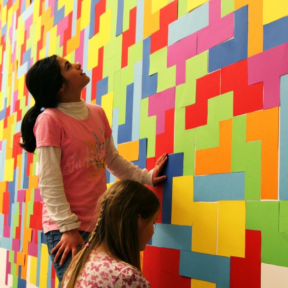 2 Mädchen vor einer mit buntem, rechteckigen Papier beklebte Wand