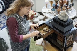 Eine Frau öffnet bei einem Schränkchen eine Schublade, im Hintergrund verschiedene Schüsseln und Pokale