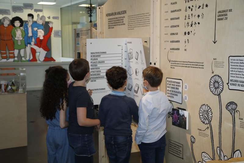 Ein Mädchen und drei Buben betrachten in der Ausstellung &quot;Umwelt, Klima & Du&quot; die Bauanleitung für ein Herbarium.