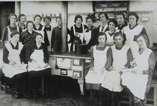 altes Foto: Hauswirtschaftskurs an der Kochmaschine. 17 Frauen stehen um einen alten Herd versammelt und schauen in die Kamera.