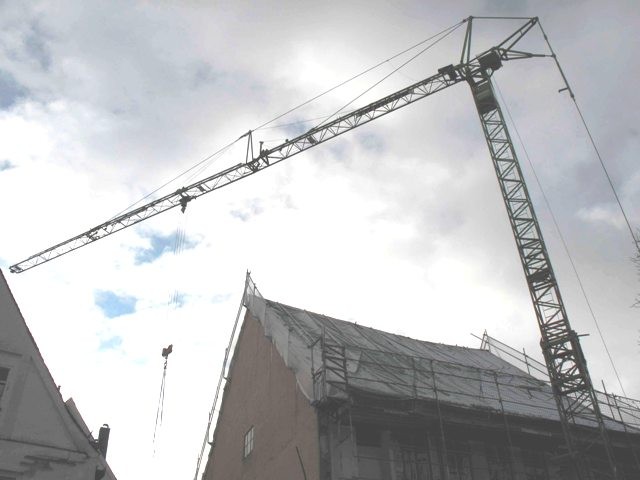 Blick in den bewölkten Himmel, im Vordergrund ein Baukran und das abgedeckte Dach eines Hauses