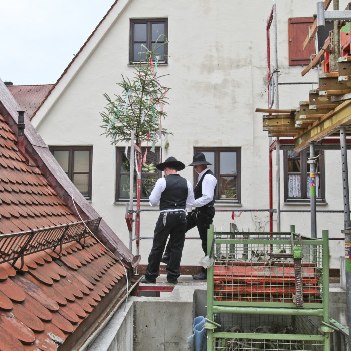 Richtfest, zwei Zimmermänner stehen auf dem Rohbau des Neubaus und verlesen den Richtspruch, links ein Gerüst auf dem verschiedene Baumaterialien liegen.