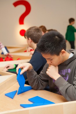 Ein Junge sitzt an einem Tisch und knobelt mit blauen Formen, dahinter mehrere Kinder, an der Wand ein großes rotes Fragezeichen