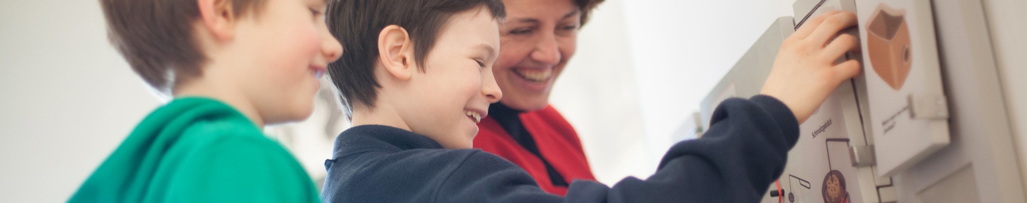 Zwei amüsierte Jungen beschäftigen sich mit einer Kinderstation. Eine Frau schaut ihnen dabei über die Schulter. 