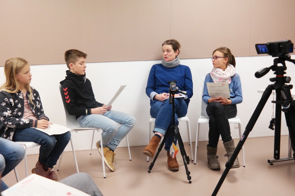 drei Kinder im Gespräch mit Nina Lutz in einem Stuhl-Halbkreis und werden von einer Kamera gefilmt