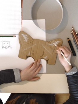 Kind malt Tiermaske mit Holzstiften an, daneben ist die Skizze eines Tigers 