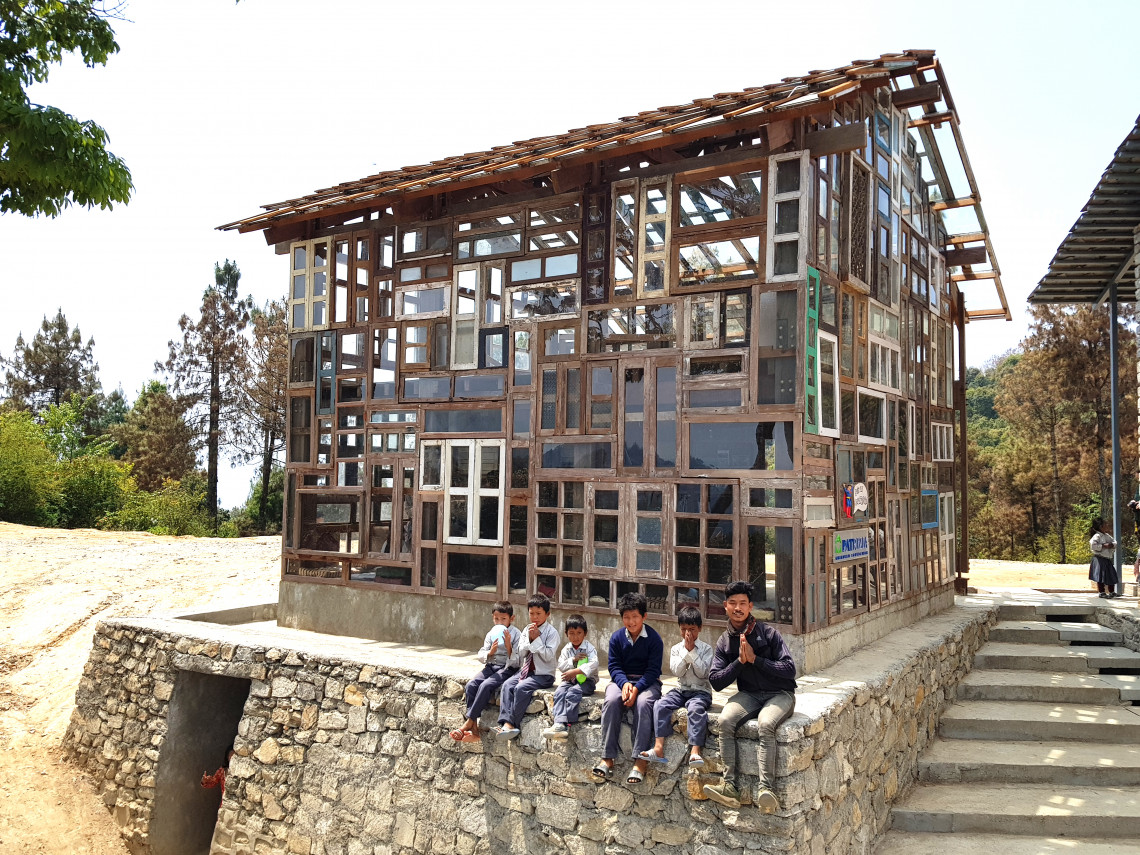 5 Kinder und ein Erwachsener sitzen auf einer Mauer vor einem Haus mit vielen verschieden gro&szlig;en Fenstern