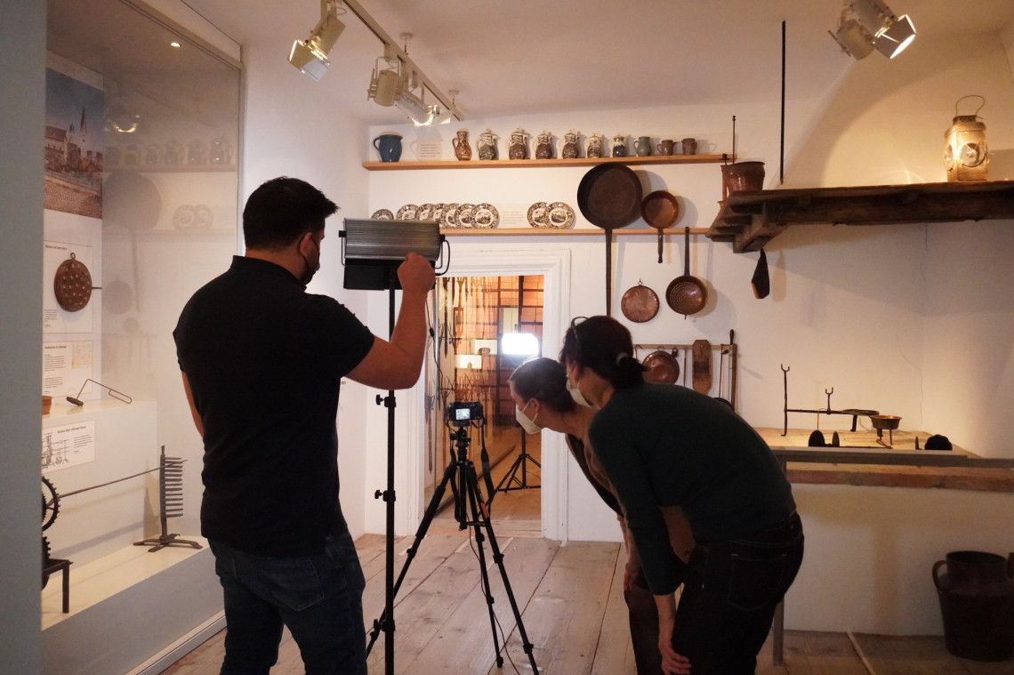 drei Teilnehmer des Technikworkshops im Ausstellungsraum Küche des Stadtmuseums Kaufbeuren, eine Person leuchtet den Raum aus, zwei Personen schauen in eine Kamera auf einem Stativ