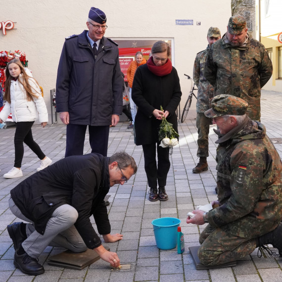 Bürgermeister Oliver Schill kniet in der Fußgängerzone und putzt einen Stolperstein, vor ihm ein hellblauer Eimer und eine Flasche Putzmittel. Weiters vier Vertreter der Bundeswehr und Museumsleiterin Petra Weber