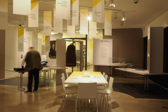 Blick in die Sonderausstellung Kaufbeuren unterm Hakenkreuz, von der Decke hängen gelb/weiße Fahnen mit Zitaten, rechts ein Besucher, welcher sich ein Objekt anschaut, mittig ein Tisch, links Ausstellungsobjekte