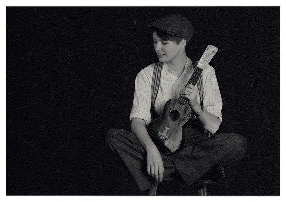 Schwarz/weiß-Foto, eine junge Frau, sitzend, gekleidet im Stil der 1920er Jahre, hält eine Ukulele in der linken Hand