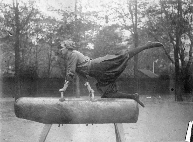 Altes schwarz/weiß-Foto einer jungen Dame, welche auf einem Pferd (Turngerät) turnt. Das Pferd steht im Freien, im Hintergrund ein Zaun, dahinter Bäume.