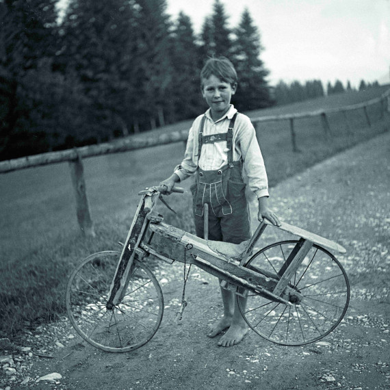 ca. fünfjähriger Junge in Lederhose, barfuß auf einem Feldweg mit einem selbstgebautem Fahrrad