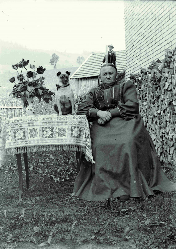 Alte Frau sitzt draußen am Tisch, auf dem neben einer Blumenvase ein Rehpinscher sitzt. Hinter ihrem Kopf fährt ein Radfahrer.